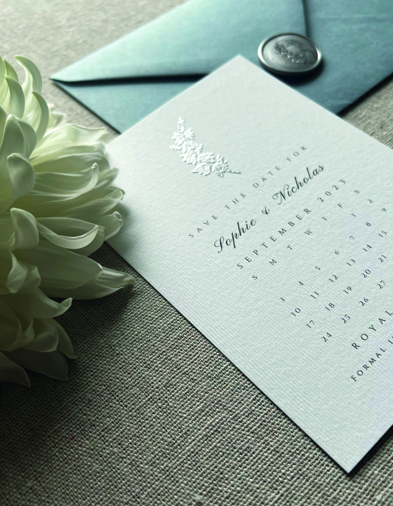 Silver embossed sprig on white save the date with white flower in foreground and blue envelope with silver wax seal